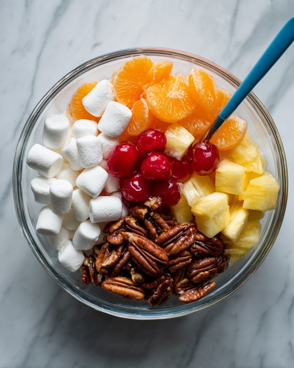 A clear glass bowl on a white marbled surface holds a layered fruit salad with visible sections of small white marshmallows, bright orange mandarin slices on top, red maraschino cherries, golden yellow pineapple chunks, and a pile of dark brown pecan halves. A blue spoon rests inside the bowl, partially submerged under the ingredients. The colors are bright and vibrant, showing a mix of textures from soft marshmallows to juicy fruit and crunchy nuts. photo taken with an iphone --ar 4:5 --v 7