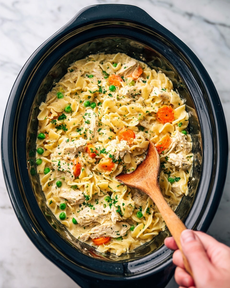 A black slow cooker bowl filled with a thick creamy pasta dish that has three visible layers: the creamy yellow sauce evenly coats the soft short noodles, chunks of white cooked chicken mixed throughout, and orange carrot slices and green peas spread within the sauce. Fresh green chopped herbs are sprinkled on top for a touch of color. A wooden spoon is inserted in the center, slightly lifting some of the pasta, and a woman's hand grips the handle. The background is a white marbled texture photo taken with an iphone --ar 4:5 --v 7