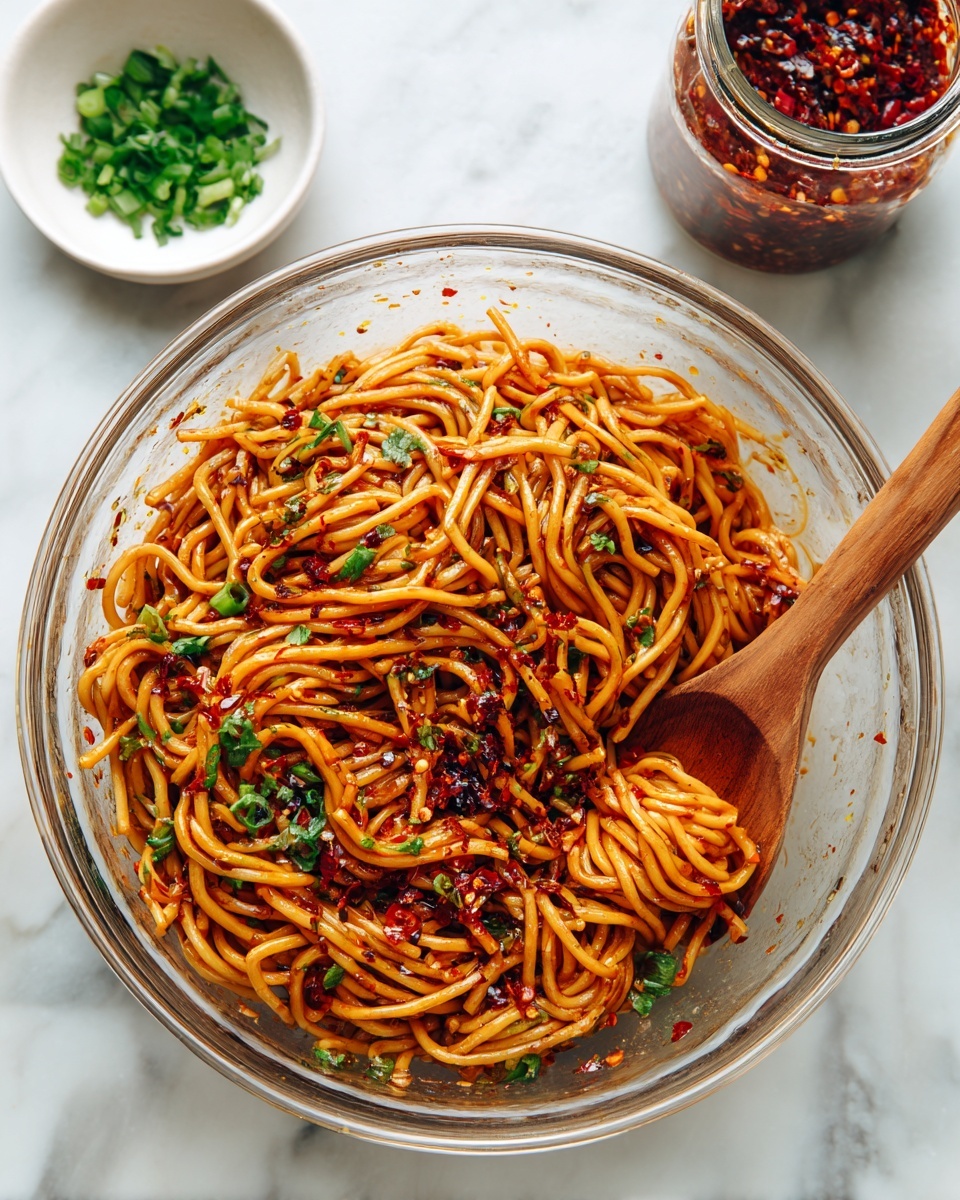 A clear glass bowl filled with about two layers of orange-brown cooked noodles mixed with dark red chili flakes and bits of green herbs scattered on top, giving a fresh and spicy look; a wooden spoon is partially buried in the noodles on the right side, showing some noodles clinging to it, all placed on a white marbled surface with a small white bowl of chopped green herbs and a jar of red chili paste nearby in the background, the photo taken with an iphone --ar 4:5 --v 7