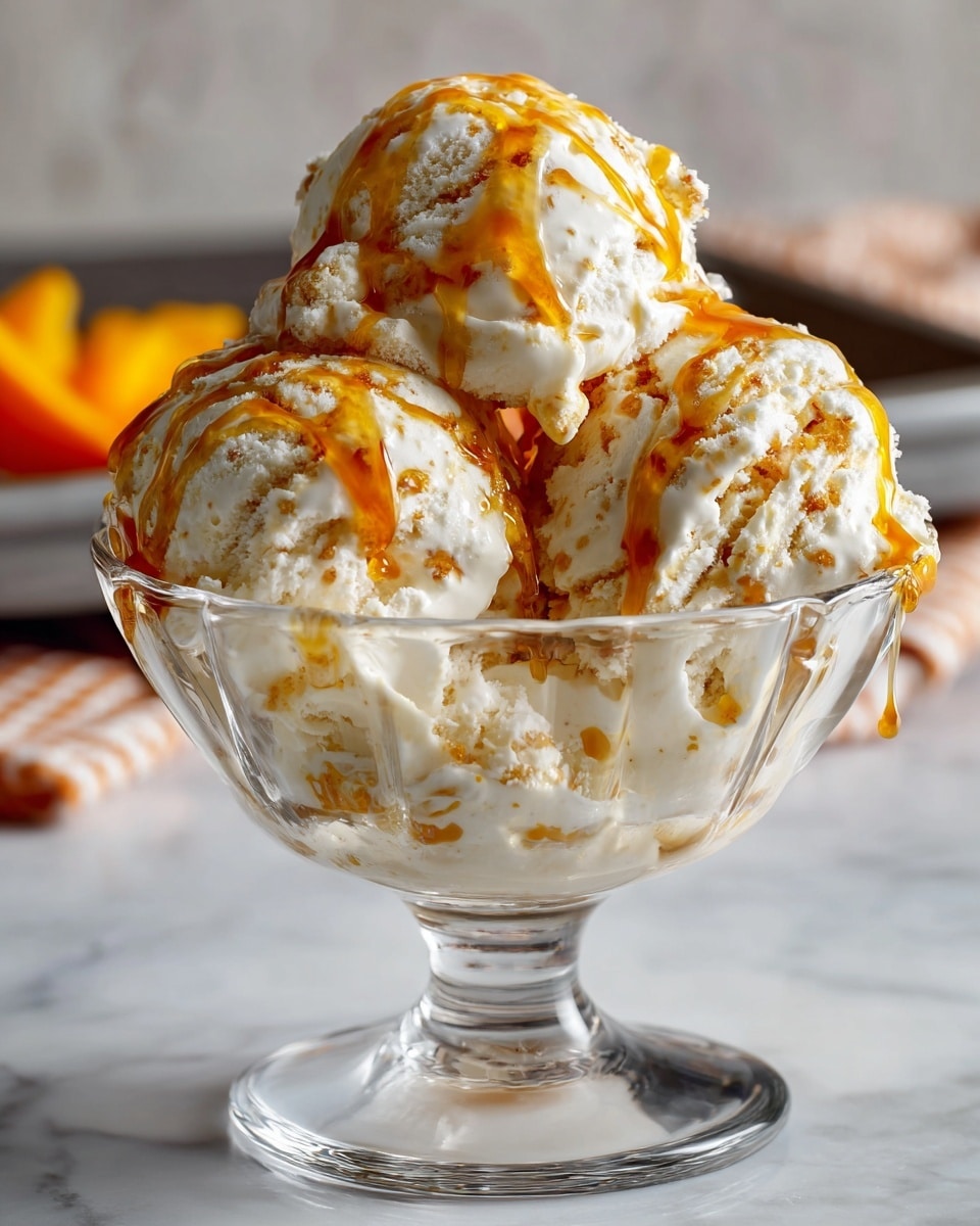 A clear glass dessert bowl with a short stem is filled with three large scoops of creamy white ice cream mixed with light brown bits, showing a rough texture. The ice cream is topped with shiny golden caramel syrup that drips down the sides. The bowl is set on a white marbled surface, with an out-of-focus gray tray and some orange-colored fruit pieces in the background. Photo taken with an iphone --ar 4:5 --v 7