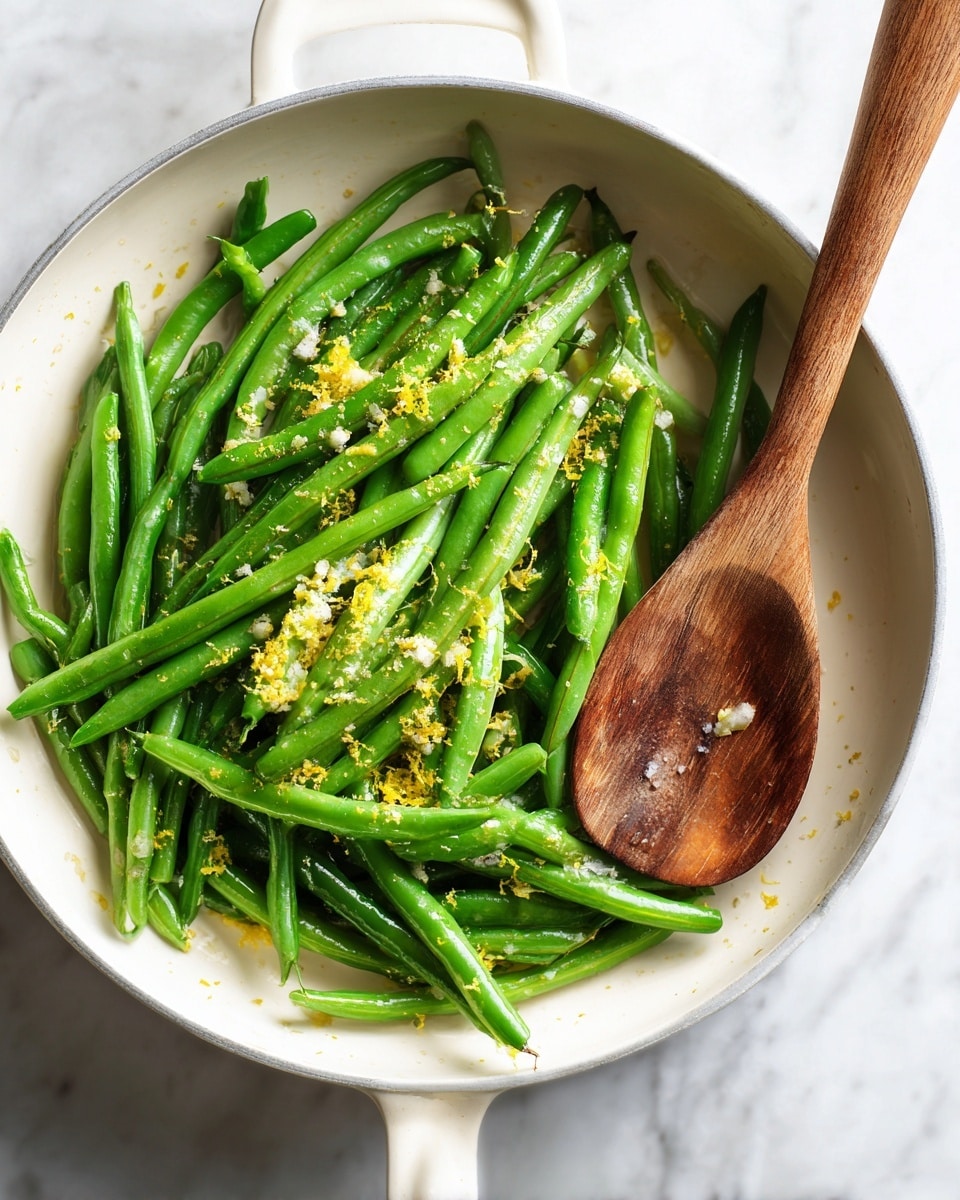 Inside a white pan, there is a single layer of bright green cooked green beans scattered around. Small bits of yellow lemon zest and light salt grains are sprinkled evenly on top, adding texture and contrast. A wooden spoon with a smooth, worn surface rests on the right edge of the pan over a white marbled surface background. The photo taken with an iphone --ar 4:5 --v 7