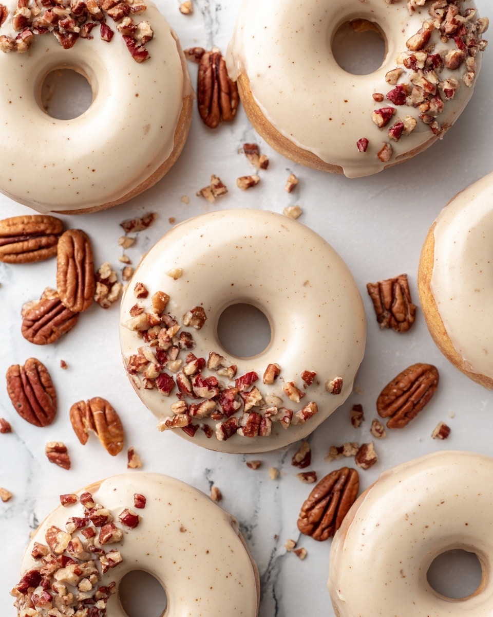 Several doughnuts are placed on a white marbled surface covered with parchment paper. Each doughnut has one layer of smooth, light beige icing covering the whole top, with a scattering of chopped pecans on one side, adding rough texture and reddish-brown color contrast. The doughnuts vary in size, with some larger and some smaller, all round with a hole in the center. Whole pecans are scattered among the doughnuts on the parchment paper, enhancing the visual appeal. photo taken with an iphone --ar 4:5 --v 7