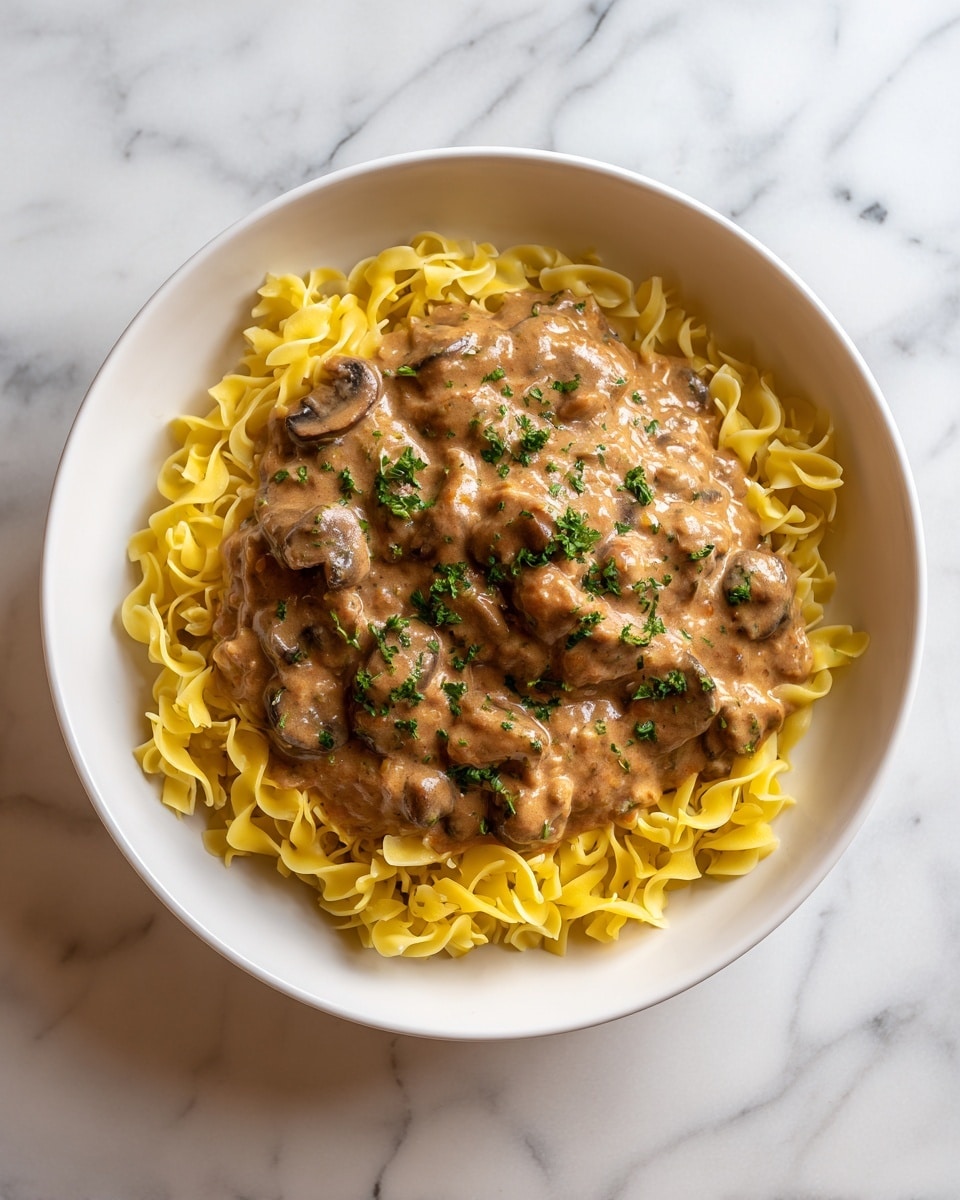A white bowl holds two main layers: the bottom layer is a bed of twisted yellow egg noodles with a smooth, soft texture, loosely arranged around the edges; on top, a thick, creamy brown sauce with visible chunks of meat and mushrooms covers the noodles mostly in the center, with small green parsley pieces sprinkled over the sauce for color contrast. The bowl sits on a white marbled surface. photo taken with an iphone --ar 4:5 --v 7