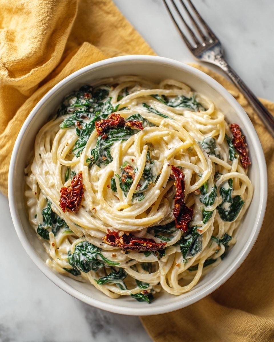 A white bowl filled with creamy spaghetti pasta, the noodles coated in a smooth white sauce. Mixed in the pasta are dark green spinach leaves and pieces of reddish-brown sun-dried tomatoes, all covered lightly in sauce. The bowl sits on a soft yellow cloth on a white marbled surface, with a silver fork resting nearby. The scene has soft, natural lighting. photo taken with an iphone --ar 4:5 --v 7