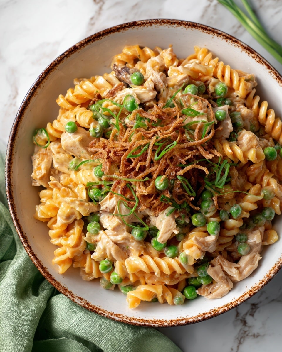 The image shows a bowl of rotini pasta dish with three visible layers: the bottom layer consists of orange rotini pasta coated in a creamy sauce, the middle layer has light brown cooked chicken pieces and bright green peas, and the top layer is garnished with fresh sliced green onions and thin, crispy fried onions, all served in a white bowl with a rustic brown rim, placed on a white marbled surface with a soft green cloth partly visible on the side photo taken with an iphone --ar 4:5 --v 7
