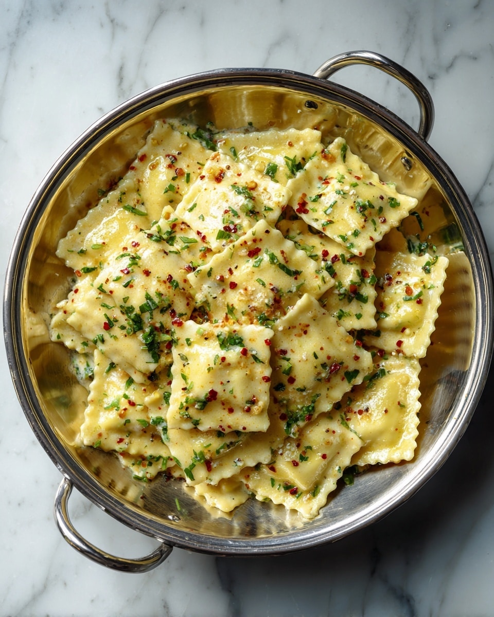 A large silver metal bowl contains many small square ravioli pieces, all light golden yellow in color with slightly browned spots. On top of the ravioli is a creamy sauce with chopped green herbs and red pepper flakes sprinkled over it. The bowl is placed on a surface with a white marbled texture. photo taken with an iphone --ar 4:5 --v 7