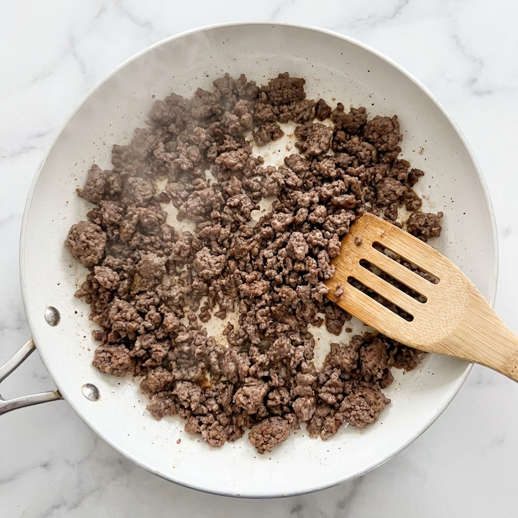 This image shows browned ground beef cooking in a white pan. The beef is broken into small, crumbly pieces that have a rich brown color with some areas darker from cooking. A light steam is rising from the beef, giving a sense of heat. A light wooden spatula with holes is partially under the beef on the right side, stirring the meat. The background is a white marbled surface. photo taken with an iphone --ar 4:5 --v 7