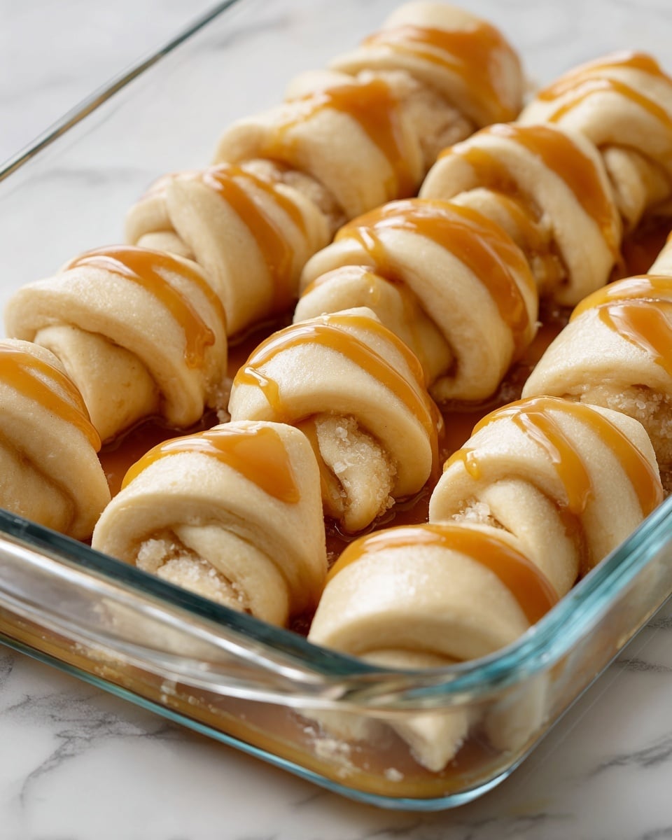 In a clear glass baking dish set on a white marbled surface, there are several small crescent rolls arranged in rows. Each roll is pale beige with a soft, doughy texture and has a light golden caramel sauce drizzled unevenly over the top. The caramel adds a glossy, sticky layer contrasting with the matte dough. The dish shows some bubbling white butter or sugar melting beneath the rolls, creating a shiny layer at the base. The rolls are closely placed but not touching, revealing the jagged, folded edges where the dough overlaps. photo taken with an iphone --ar 4:5 --v 7