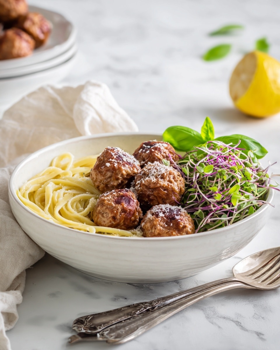 A white bowl on a white marbled surface holds a layered dish starting with a base of light yellow pasta on the left side, topped with five round, browned meatballs that are slightly textured and sprinkled with a light dusting of white cheese. To the right of the meatballs is a small heap of fresh mixed green sprouts with thin purple stems. A bright green basil leaf sits on one of the meatballs and a vintage silver fork and spoon rest beside the bowl on the right. In the softly blurred background, there is a halved lemon and another white plate with more meatballs and pasta. photo taken with an iphone --ar 4:5 --v 7