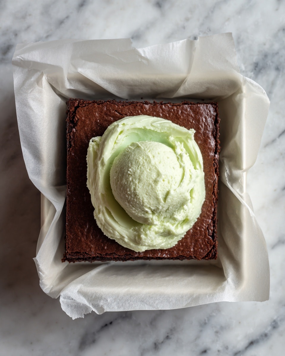 A square baking pan lined with white parchment paper holds a thick, dark brown brownie layer with a slightly cracked surface. On top of the brownie, in the middle, there is a dollop of light green frosting with a creamy, smooth texture that looks freshly spread and swirled. The pan sits on a white marbled surface. photo taken with an iphone --ar 4:5 --v 7