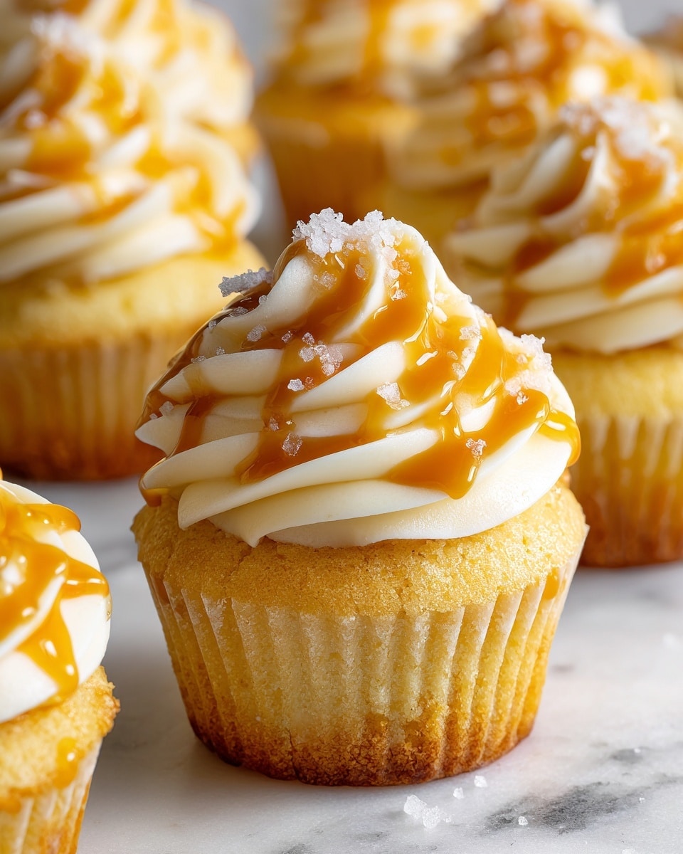 A close-up view of a single cupcake with two main layers visible: the base is a light golden-yellow cake with a soft, slightly spongy texture, topped with a large swirl of creamy, off-white frosting that has a smooth, fluffy look. The frosting is decorated with thin streams of shiny, golden caramel sauce drizzled over it, adding a glossy texture. Small flakes of coarse salt are scattered on the frosting, creating tiny white crystals that contrast with the caramel. In the blurred background, more cupcakes with similar frosting and caramel drizzles can be seen, all placed on a white marbled surface. photo taken with an iphone --ar 4:5 --v 7