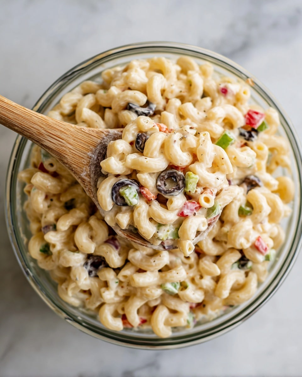 A close-up from above shows a clear round bowl filled with a creamy macaroni salad. The salad has three main layers: the pale yellow cooked macaroni shaped like curved elbows, scattered black olive slices creating dark rings, and small pieces of red bell pepper adding a bright red touch. There are also hints of green herbs and celery bits spread evenly throughout, all coated in a light creamy dressing that gives a slight shine. A wooden spoon holds a small scoop of the salad at the center, highlighting the detailed texture of the macaroni and colorful mix of olives, peppers, and herbs. The bowl rests on a white marbled surface. Photo taken with an iphone --ar 4:5 --v 7