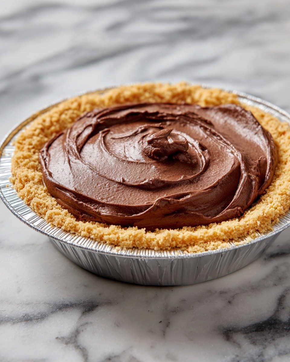 The image shows a pie with two visible layers placed on a white marbled surface. The bottom layer is a light brown, crumbly crust forming the base and sides of the pie, held in a round foil pie pan. On top of this crust is a thick, dark brown, smooth chocolate filling dolloped in the center, appearing rich and creamy with slight glossy texture. photo taken with an iphone --ar 4:5 --v 7