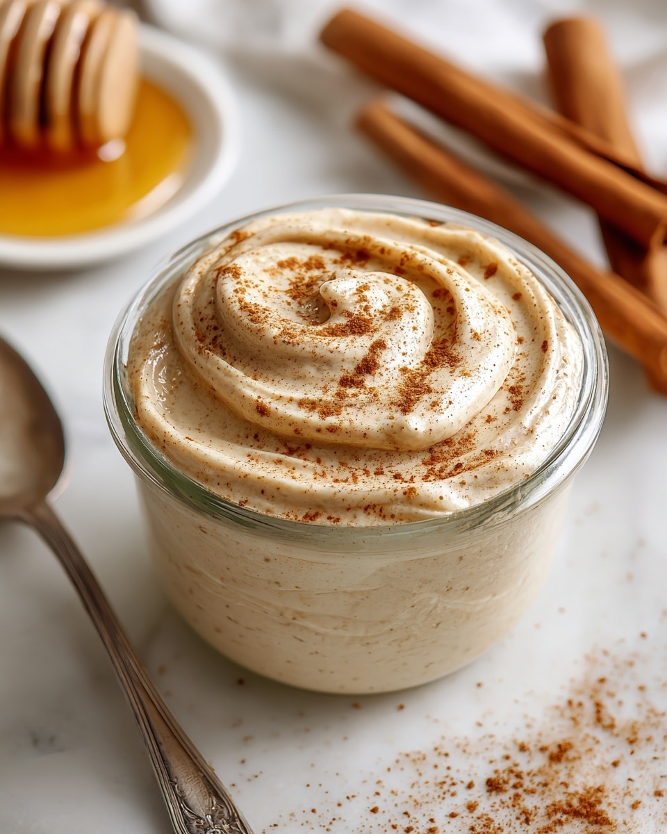 A clear glass jar filled with a smooth, creamy light beige spread that has soft swirls and ridges on the surface. Tiny brown specks of cinnamon powder are sprinkled evenly on top and around the edges of the spread, adding texture and color contrast. The jar sits on a white marbled surface with a small shiny silver spoon partially visible at the bottom left. In the background, there are blurry cinnamon sticks and a honey dipper resting on a small puddle of golden honey, all softly out of focus. Photo taken with an iphone --ar 4:5 --v 7