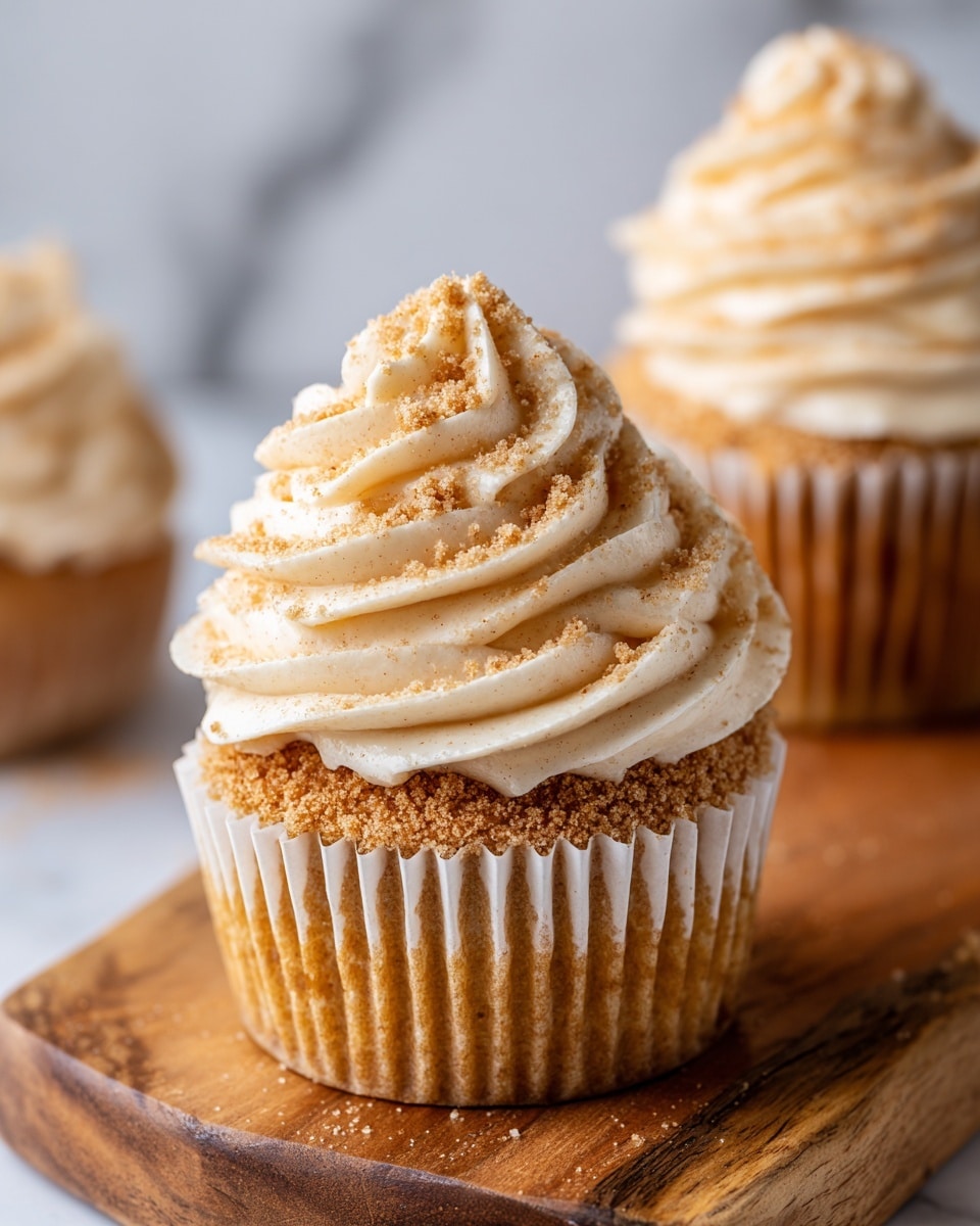 A close-up view of a single cupcake in a white paper liner on a wooden board, showing three thick swirls of creamy beige frosting dusted with light brown crumbs on top. The cupcake base is golden brown with a soft texture, and the frosting has a smooth, fluffy look with a small rounded peak in the center. Another cupcake is partly visible in the top right corner, placed on the same wooden board. The background features a white marbled surface. photo taken with an iphone --ar 4:5 --v 7