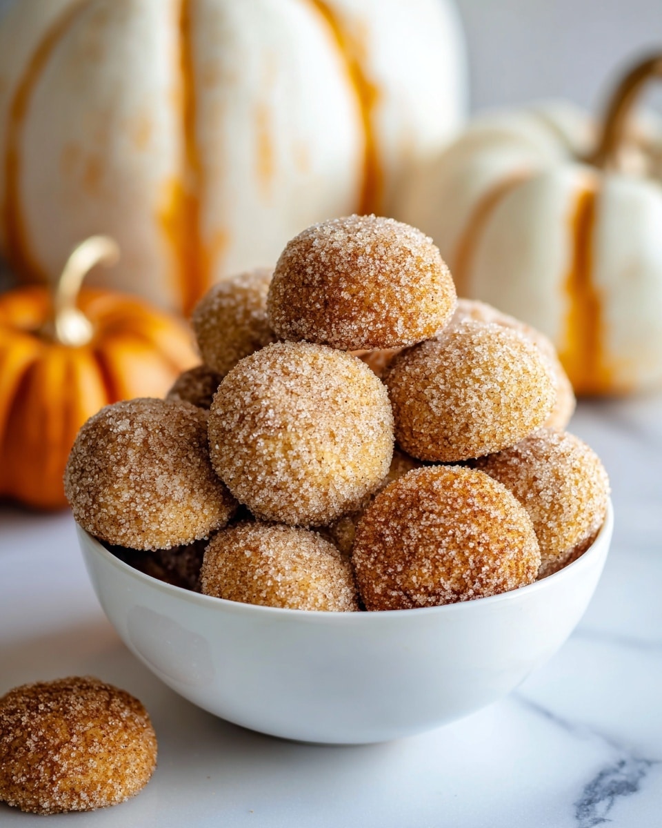 A white bowl is filled with round cookies, each covered in a textured layer of sugar mixed with cinnamon, giving them a grainy light brown and tan surface. The cookies have a slightly cracked top showing a warm orange-brown color beneath the sugar layer. The bowl is placed on a white marbled surface with a couple of similar cookies nearby and two white pumpkins with orange streaks are in the background, adding a seasonal touch. The overall look is warm and cozy. photo taken with an iphone --ar 4:5 --v 7