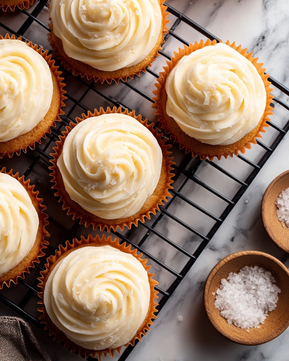 A close-up top view of six cupcakes with a single thick swirl of creamy, off-white frosting on each. The frosting has a smooth texture with tiny specks, arranged in a rosette pattern, sitting on light brown cake bases inside bright orange paper liners. The cupcakes rest on a black metal cooling rack placed over a white marbled surface. A small wooden bowl filled with coarse white salt is partly visible on the right edge of the image. photo taken with an iphone --ar 4:5 --v 7