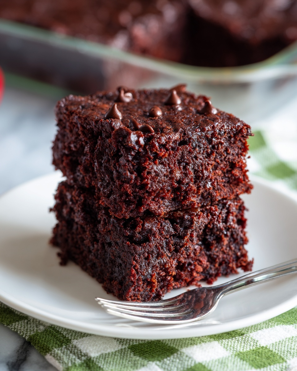 A close-up image of two square pieces of rich, dark brown chocolate cake stacked on a white plate. The cake has a slightly rough texture with visible chocolate chips scattered inside. In the background, there is a white plate holding a glass pan of the same chocolate cake on a green and white checked cloth on a white marbled surface. A silver fork lies on the plate below the cake pieces. Photo taken with an iphone --ar 4:5 --v 7