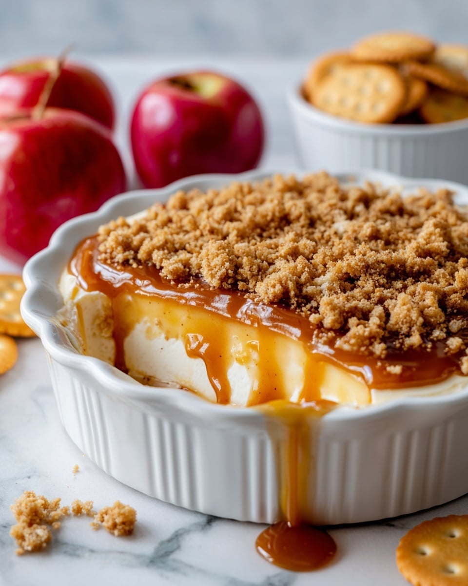 A white scalloped dish filled with a dessert showing three clear layers: the bottom layer is smooth and creamy with a pale off-white color, the middle layer is bright golden caramel sauce that drips slightly on the edges, and the top layer is crumbly with a light brown streusel texture scattered unevenly over the caramel. In the background on a white marbled surface, there are two shiny red apples and a white bowl filled with round crackers. Photo taken with an iphone --ar 4:5 --v 7
