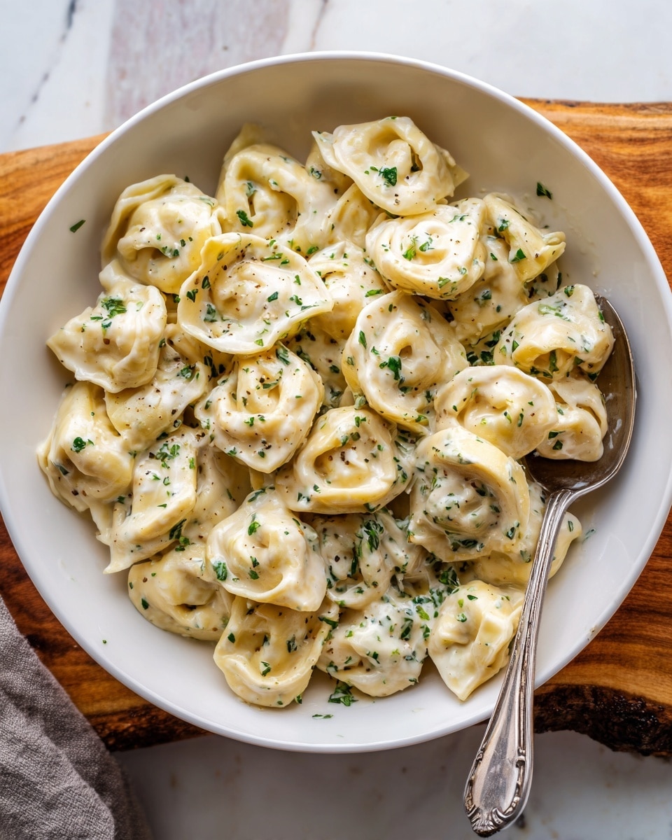 A white bowl is filled with tortellini pasta covered in a creamy white sauce with small green herb bits. The tortellini pieces are slightly folded and soft, coated evenly with the sauce that has a smooth texture with a hint of oil giving a slight shine. A silver spoon rests inside the bowl on the right side, holding some pasta pieces. The bowl is placed on a white marbled surface with a natural wood piece partly underneath it. The lighting is natural and soft, highlighting the creamy sauce and pasta shapes. photo taken with an iphone --ar 4:5 --v 7