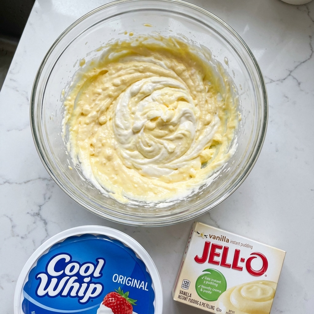 The image shows a clear glass bowl filled with a thick, creamy mixture that has a swirled texture of white and pale yellow colors spread unevenly across the surface. The mixture looks fluffy and smooth with some visible small lumps in the yellowish areas. Below the bowl, there is a round white container with a blue lid labeled