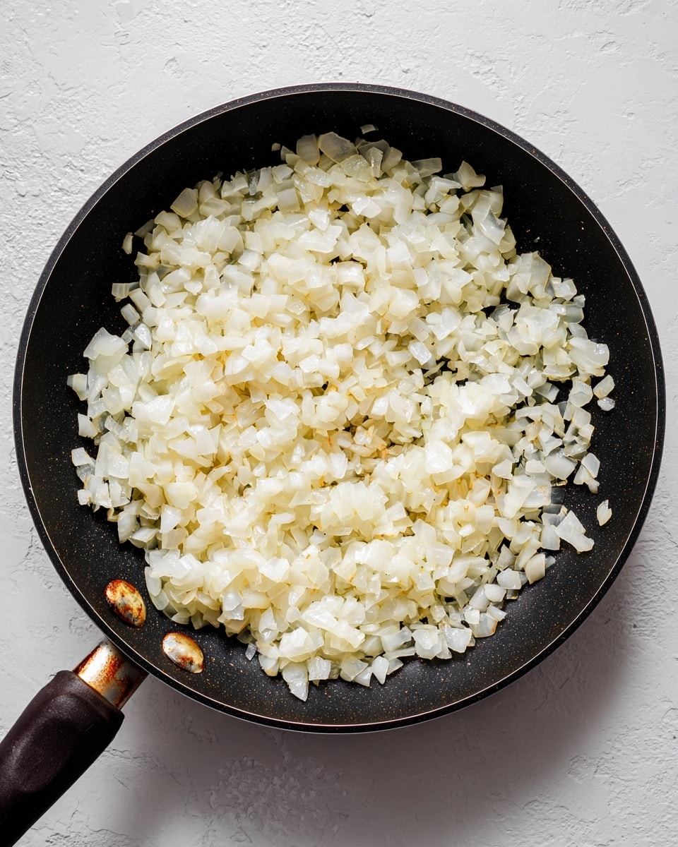A close-up view of a black frying pan filled with small, soft, cooked onion pieces that are translucent white with light golden edges, spread evenly across the pan's surface. The pan has a dark handle resting on a white marbled texture surface. The background shows a slightly stained wall that contrasts with the clean look of the pan and onions. photo taken with an iphone --ar 4:5 --v 7