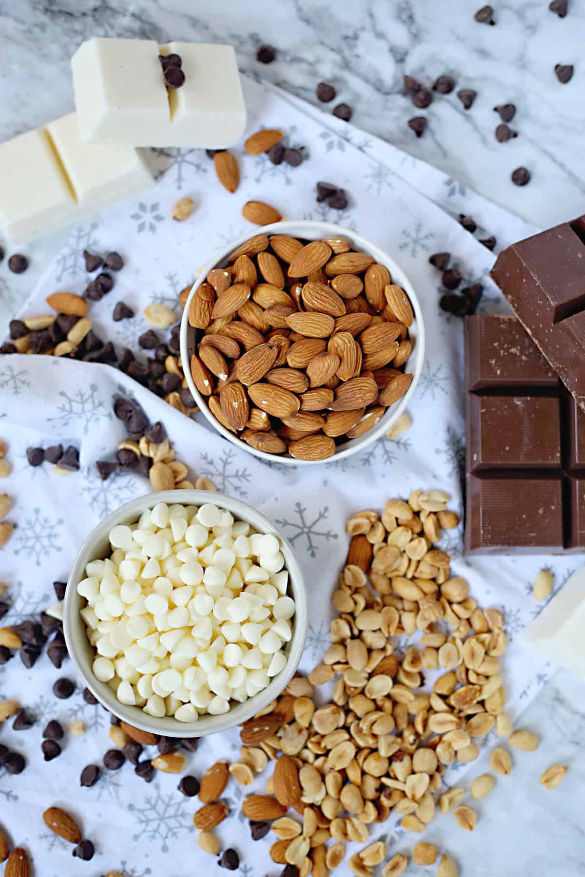 The image shows two white bowls on top of a white cloth with gray snowflake patterns, placed on a white marbled surface. The top bowl is filled with whole, brown almonds that have a smooth texture and natural shine. Below, the second bowl contains many white, round white chocolate chips with a smooth surface. Around the bowls, there are scattered roasted peanuts, dark chocolate chips, and whole almonds. To the right, there are pieces of a dark brown chocolate bar with a slightly matte finish, and to the left in the background, there are thick blocks of creamy white chocolate with a few dark chocolate chips placed on top. The overall set-up is simple and natural, with a mix of smooth and rough textures. photo taken with an iphone --ar 4:5 --v 7